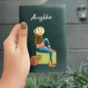 A hand holding a green personalized passport cover with a printed illustration of a woman sitting on a suitcase