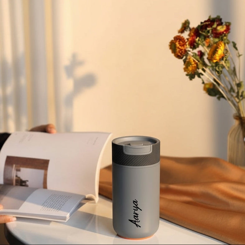 Person holding a magazine next to a gray tumbler with 'Aqua' branding on a wooden surface.